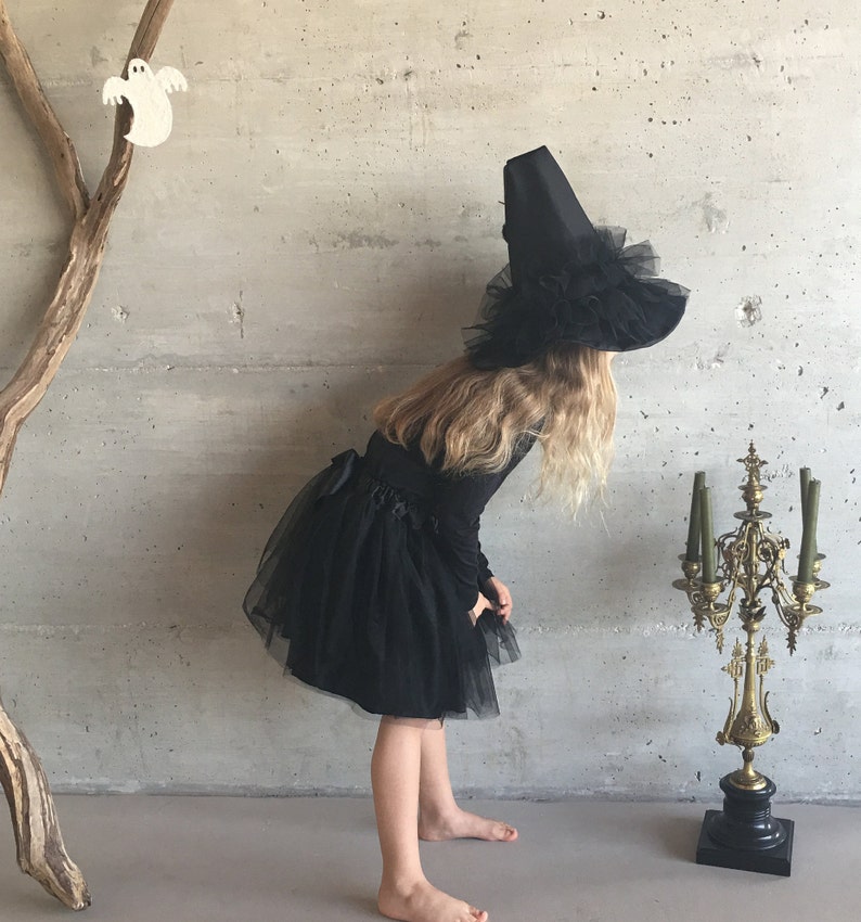 May include: A young girl dressed as a witch for Halloween. She is wearing a black tulle skirt and a black witch hat with a wide brim and a black feather trim. She is standing in front of a gray wall with a wooden branch behind her.