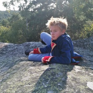May include: A person wearing a blue jacket with red and gold detailing, seated on a large rock. The jacket has a high collar and decorative buttons. The person is wearing blue jeans and black shoes. The background shows trees and a sunny sky.
