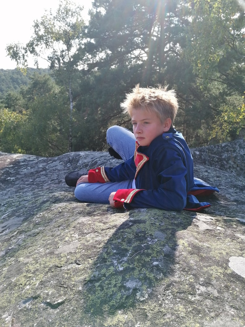 May include: A person wearing a blue jacket with red and gold detailing, seated on a large rock. The jacket has a high collar and decorative buttons. The person is wearing blue jeans and black shoes. The background shows trees and a sunny sky.