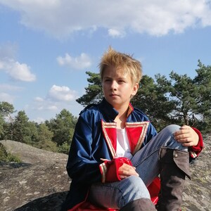 May include: A person wearing a blue, red, and gold costume coat, white shirt, and blue jeans sits on a rock. The coat has gold trim and red cuffs. The background includes trees and a blue sky with clouds.