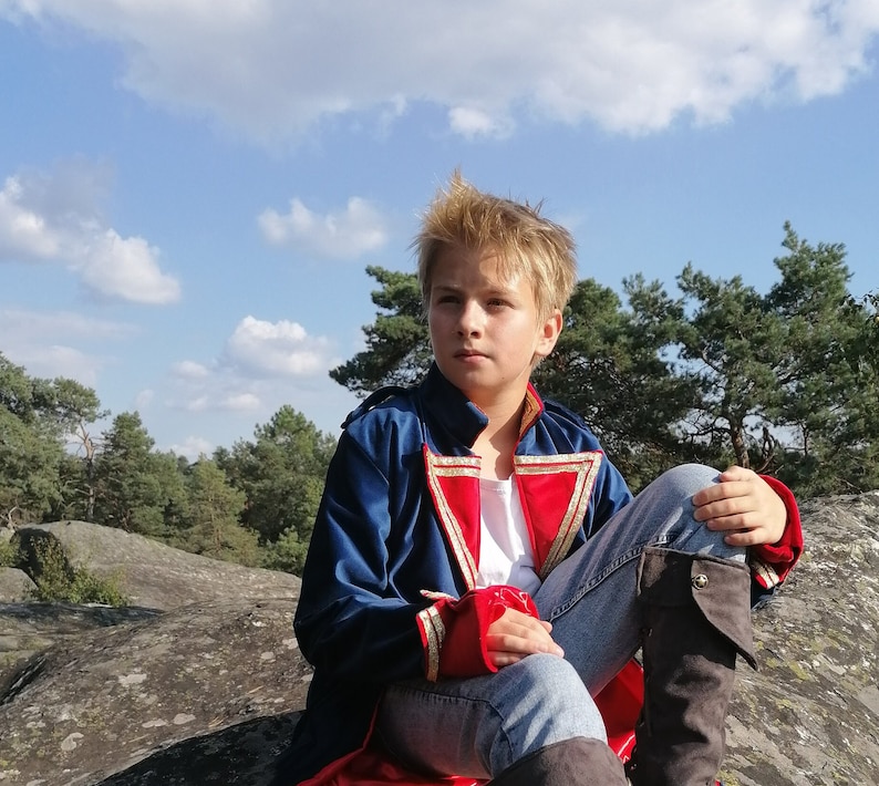 May include: A person wearing a blue, red, and gold costume coat, white shirt, and blue jeans sits on a rock. The coat has gold trim and red cuffs. The background includes trees and a blue sky with clouds.