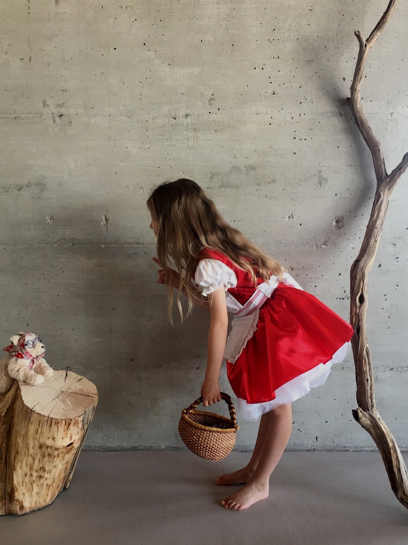 May include: A child dressed in a Little Red Riding Hood costume, featuring a red dress with a white apron and sleeves, and a woven basket. A stuffed bear wearing glasses and a scarf sits on a wooden stump.