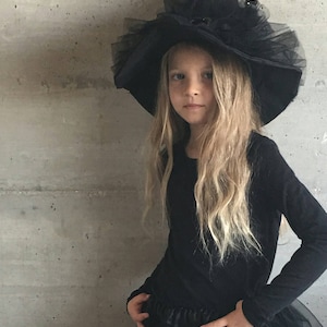 May include: A young girl dressed as a witch for Halloween. She is wearing a black witch hat with a tulle trim and a black dress.