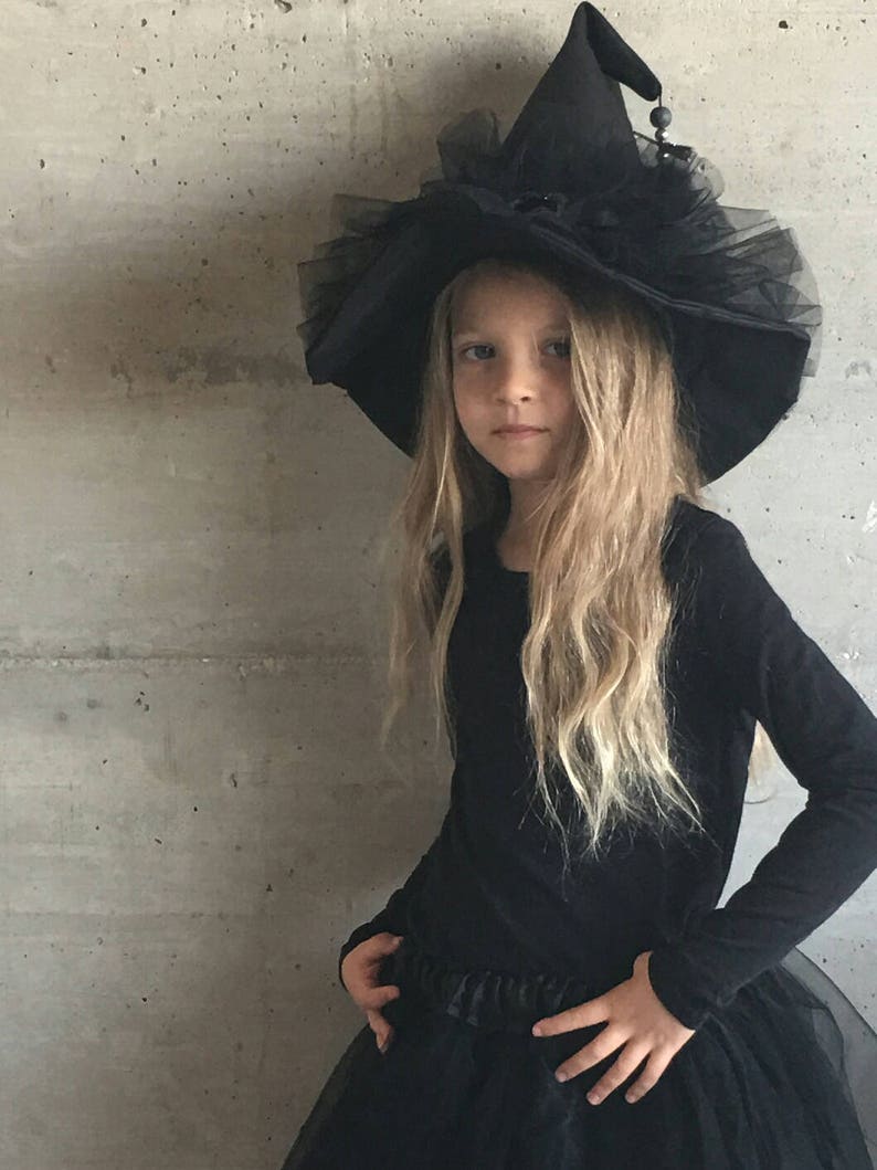 May include: A young girl dressed as a witch for Halloween. She is wearing a black witch hat with a tulle trim and a black dress.