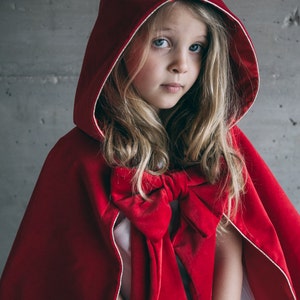 May include: A red velvet hooded cape with a large bow. The cape has a white trim around the hood and is worn by a person with blonde hair and blue eyes. The cape is a costume.