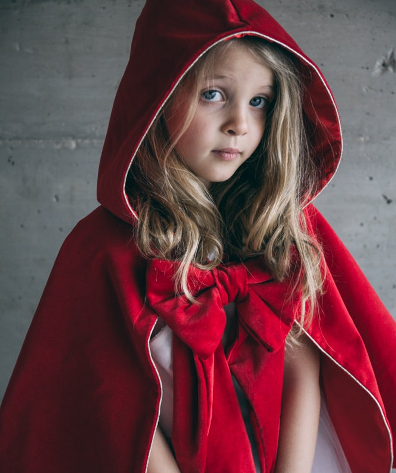 May include: A red velvet hooded cape with a large bow. The cape has a white trim around the hood and is worn by a person with blonde hair and blue eyes. The cape is a costume.