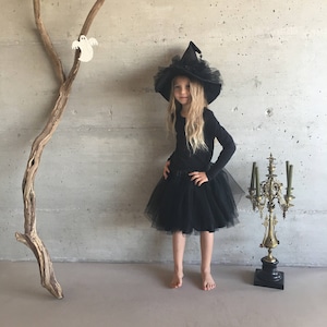 May include: A young girl dressed as a witch for Halloween. She is wearing a black tulle skirt, a black long-sleeved shirt, and a black witch hat with a tulle brim. She is standing in front of a gray wall with a wooden branch and white ghost decorations.