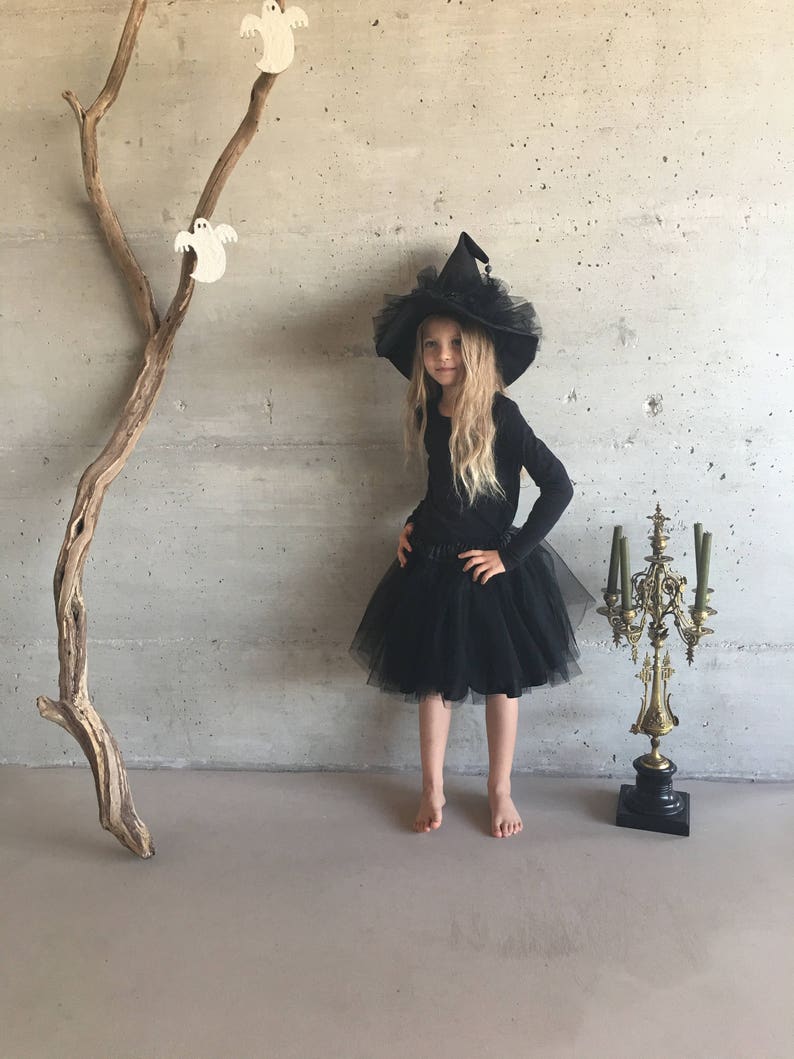 May include: A young girl dressed as a witch for Halloween. She is wearing a black tulle skirt, a black long-sleeved shirt, and a black witch hat with a tulle brim. She is standing in front of a gray wall with a wooden branch and white ghost decorations.