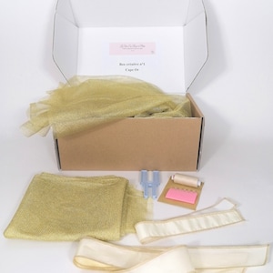Può includere: Una scatola artigianale aperta, etichettata "Box créative n°1 Cape Or", contiene tessuto a rete dorato, nastro color crema, filo e accessori per cucire. Il contenuto è disposto su una superficie bianca.