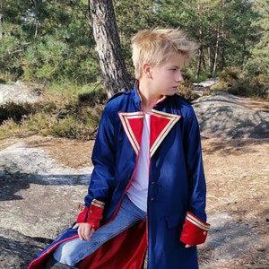 May include: A long, navy blue coat with red lining and gold trim. The coat has red cuffs and a high collar. The coat is worn over a white shirt and blue jeans. The coat is buttoned up the front.