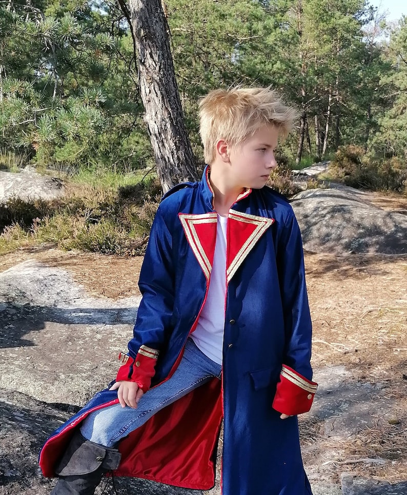 May include: A long, navy blue coat with red lining and gold trim. The coat has red cuffs and a high collar. The coat is worn over a white shirt and blue jeans. The coat is buttoned up the front.