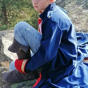 May include: A person wearing a navy blue coat with red trim and gold buttons, blue jeans, and brown boots. The coat has a high collar and a military style. The person is sitting on a rock outdoors.