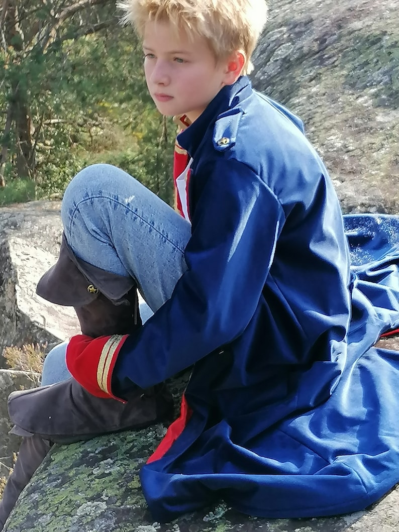 May include: A person wearing a navy blue coat with red trim and gold buttons, blue jeans, and brown boots. The coat has a high collar and a military style. The person is sitting on a rock outdoors.