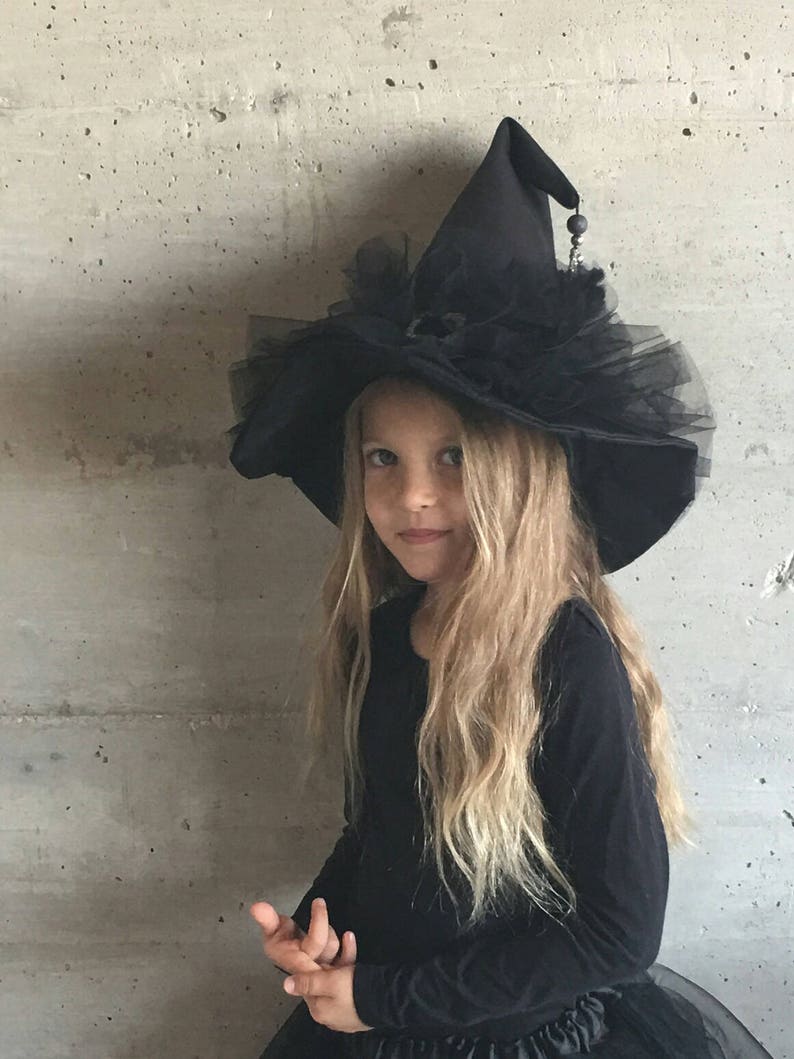 May include: A black witch hat with a wide brim, black tulle, and a silver bead accent. The hat is worn by a child with long blonde hair.
