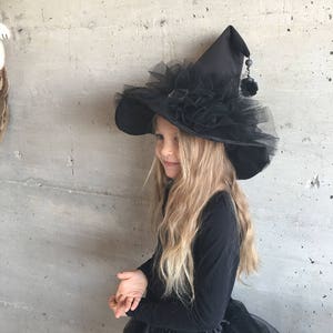 May include: A black witch hat with a wide brim, black tulle, and a black pom-pom on the top. The hat is worn by a child with long blonde hair.