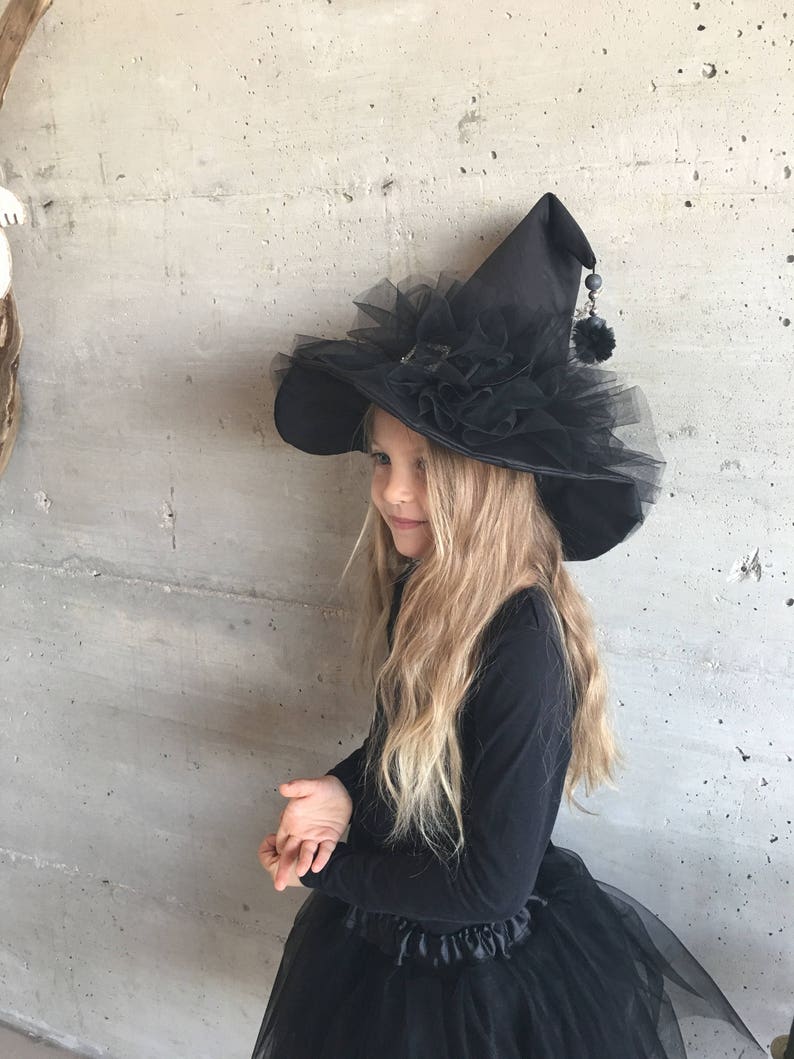 May include: A black witch hat with a wide brim, black tulle, and a black pom-pom on the top. The hat is worn by a child with long blonde hair.
