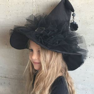 May include: A black witch hat with a large brim and a tulle ruffle around the crown. The hat has a black pom-pom and a beaded chain hanging from the tip.