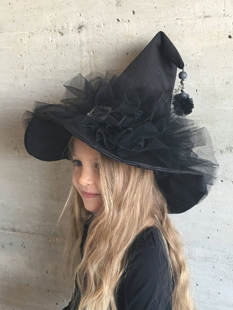 May include: A black witch hat with a large brim and a tulle ruffle around the crown. The hat has a black pom-pom and a beaded chain hanging from the tip.