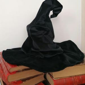 May include: A black velvet witch's hat sits atop a stack of vintage books. The hat has a tall, pointed crown and a wide brim.