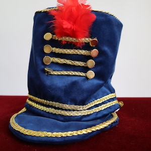Puede incluir: Un sombrero de terciopelo azul con ribete de trenza dorada y una pluma roja en la parte superior. El sombrero tiene cinco botones dorados en la parte delantera.