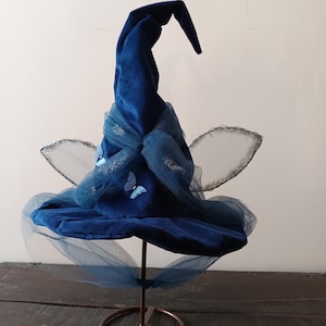 May include: A dark blue velvet witch hat with a pointed tip, decorated with sheer blue fabric, glitter, and butterfly details. The hat is displayed on a bronze stand, with silver fairy wings attached.
