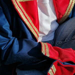 May include: A blue jacket with red and gold detailing. The jacket has red triangular panels on the shoulders and cuffs, edged with gold trim. The sleeves are blue, and it's worn over a white top and blue jeans. The gold trim adds a touch of elegance.