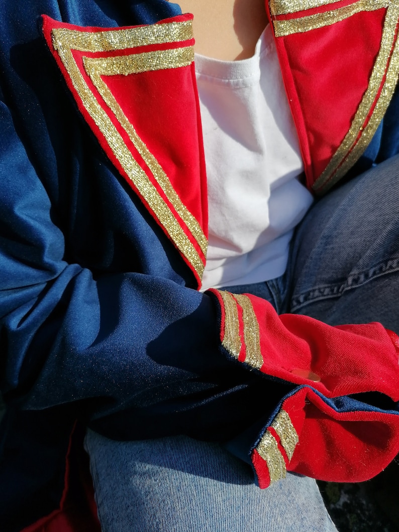 May include: A blue jacket with red and gold detailing. The jacket has red triangular panels on the shoulders and cuffs, edged with gold trim. The sleeves are blue, and it's worn over a white top and blue jeans. The gold trim adds a touch of elegance.