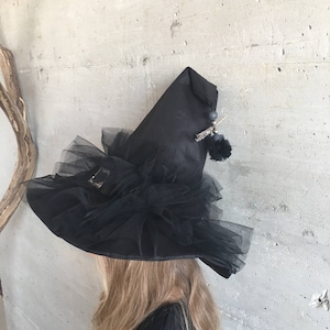 May include: A black witch hat with a wide brim and a tall pointed crown. The hat is decorated with black tulle and a small black pom-pom.