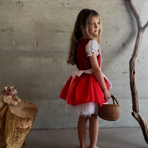 May include: A red Little Red Riding Hood costume with white sleeves and a white underskirt. The costume includes a matching red skirt and a woven basket. The girl is standing barefoot.