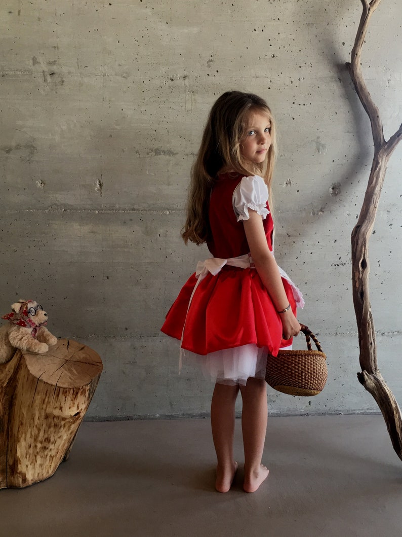 May include: A red Little Red Riding Hood costume with white sleeves and a white underskirt. The costume includes a matching red skirt and a woven basket. The girl is standing barefoot.
