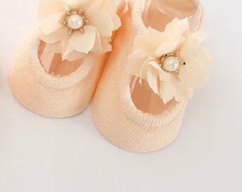 Könnte beinhalten: Ein Paar beige Baby-Socken mit einer zarten Blumenverzierung und einem Perlenakzent.