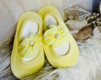 Op de afbeelding: Een paar gele babysokken met een tule strik en parelversiering. De sokken liggen op een wit, pluche oppervlak.