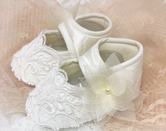 Könnte beinhalten: Ein Paar weiße Babyschuhe mit Spitzendetails und einer zarten Blumenverzierung.