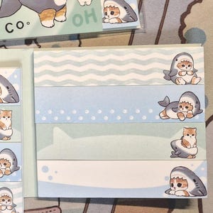 May include: Several packs of Mofusand cat-themed sticky notes. The notes feature illustrations of cats dressed as sharks. The color palette includes blue, white, and tan. The text "mofusand" is visible on the packaging.