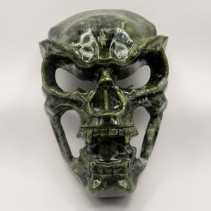 May include: A green jade skull mask with detailed carvings. The mask has a menacing expression with a wide open mouth and sharp teeth.