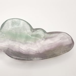 May include: A cloud-shaped decorative dish made of translucent fluorite. The dish features a blend of colors, including lavender, green, and gray. The smooth, polished surface reflects light, highlighting the natural patterns within the stone.