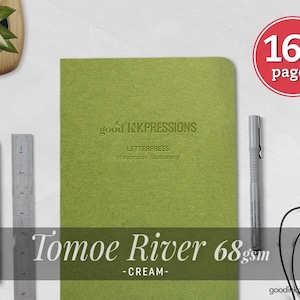 May include: A green notebook with the embossed text good INKPRESSIONS LETTERPRESS Handmade Stationery. A red circle graphic states 160 pages. A metal ruler, a black pencil, a grey pen, and black wired earbuds are positioned nearby.