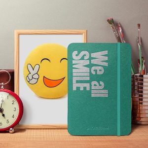 Può includere: Un diario verde con il testo "We all smile" in lettere argentate. Il diario ha un segnalibro a nastro argentato ed è appoggiato su una superficie di legno accanto a un orologio e una cornice con un cuscino giallo a forma di smiley.