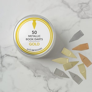 Puede incluir: Una lata redonda dorada con el texto "50 Metallic Book Darts GOLD" y la marca "good inkpressions". Varias flechas metálicas para libros en plata, oro y cobre están dispersas alrededor de la lata sobre una superficie de mármol.