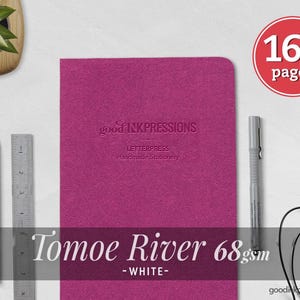 Puede incluir: Un cuaderno magenta con el texto en relieve good INKPRESSIONS LETTERPRESS Handmade Stationery. Una insignia circular roja indica 160 páginas. Un lápiz negro, una regla de metal, un bolígrafo gris y auriculares con cable negros están colocados cerca.