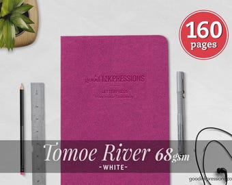 May include: A magenta notebook with the embossed text good INKPRESSIONS LETTERPRESS Handmade Stationery. A red circular badge indicates 160 pages. A black pencil, a metal ruler, a gray pen, and black wired earbuds are positioned nearby.