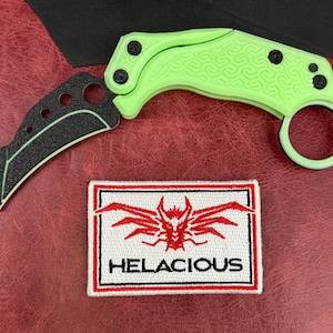 May include: A neon green karambit knife with a black blade and a ring handle. The knife is next to a rectangular patch with the word "HELACIOUS" and a red graphic.