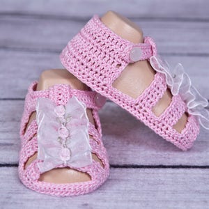 Puede incluir: Un par de sandalias rosas de bebé hechas a ganchillo. Las sandalias tienen un cierre de botón y están adornadas con una cinta blanca y flores. Las sandalias se muestran sobre una superficie de madera clara.