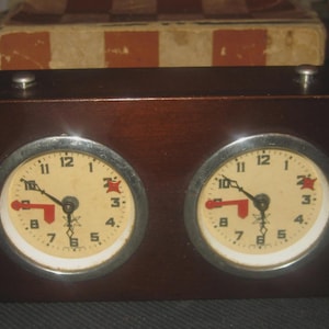 May include: A rectangular dark brown wooden analog chess clock featuring two circular clock faces with cream-colored dials, black numerals, and silver-toned bezels. Each clock face includes black hour and minute hands and a red indicator hand.