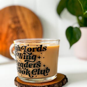 May include: A glass mug filled with a light brown drink, set on a wooden coaster. The mug has black text saying "High Lords Wing Leaders Book Club" with star details. A wooden board and a potted plant are in the background.