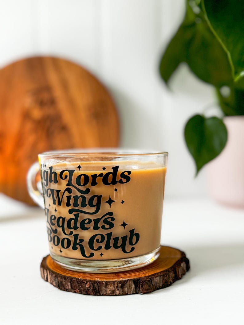 May include: A glass mug filled with a light brown drink, set on a wooden coaster. The mug has black text saying "High Lords Wing Leaders Book Club" with star details. A wooden board and a potted plant are in the background.