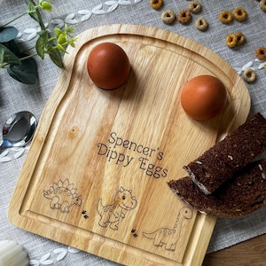 May include: Wooden bread-shaped serving board with the text "Spencer's Dippy Eggs" and dinosaur illustrations. Two brown eggs and slices of dark bread are on the board. Cereal pieces and a spoon are in the background.