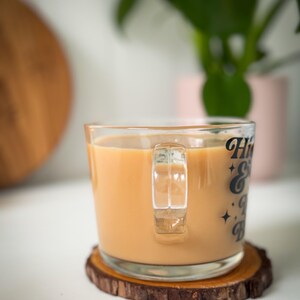 May include: A glass mug containing a light brown beverage, resting on a wooden coaster. The mug features the text "Hi & Bye" in black. A green plant is in the background. The coaster is made of a slice of wood.