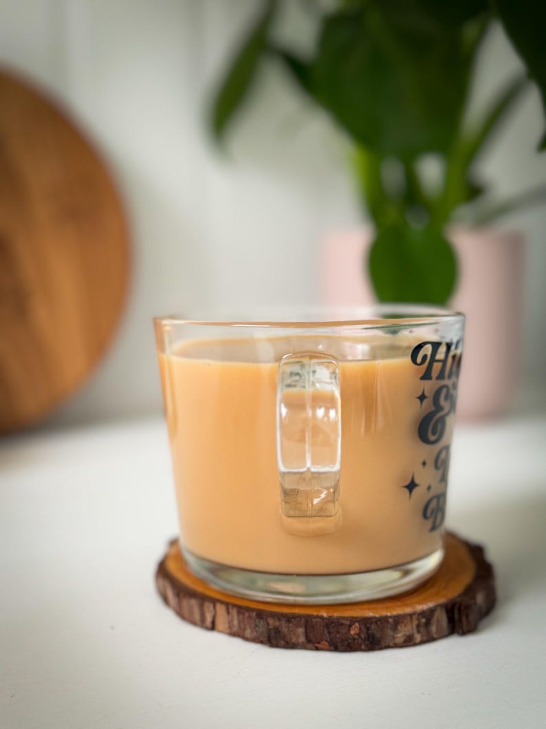 May include: A glass mug containing a light brown beverage, resting on a wooden coaster. The mug features the text "Hi & Bye" in black. A green plant is in the background. The coaster is made of a slice of wood.