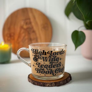 May include: A clear glass mug filled with a light brown drink, inscribed with "High Lords & Wing Leaders Book Club" in black lettering. The mug rests on a small, round wooden coaster. A lit green candle and a wooden board are in the background.